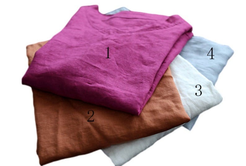 May include: Four folded linen shirts in different colors: a deep magenta, a warm brown, a soft white, and a light blue. Each shirt has a simple, round neckline. The shirts are stacked on top of each other, showcasing the variety of colors.