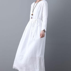 May include: A white linen dress with long sleeves and a drawstring waist. The dress has a v-neckline and a relaxed fit.