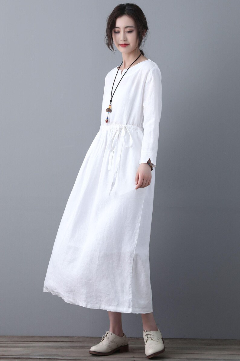 May include: A white linen dress with long sleeves and a drawstring waist. The dress has a v-neckline and a relaxed fit.