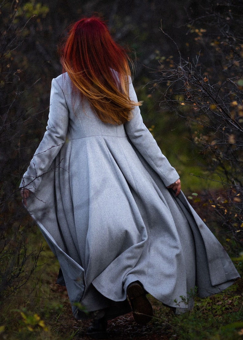 May include: A long, light gray coat with a flared skirt is worn by a person walking away from the camera. The person has long red hair with a gradient to blonde. The coat is made of a soft material and is suitable for fall or winter.
