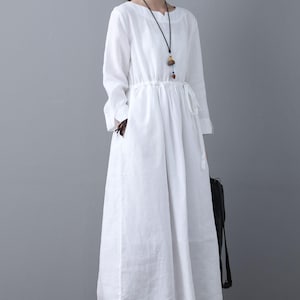 May include: A white linen dress with long sleeves and a drawstring waist. The dress has a relaxed fit and a slightly flared skirt. The model is wearing the dress with a simple necklace and white shoes.