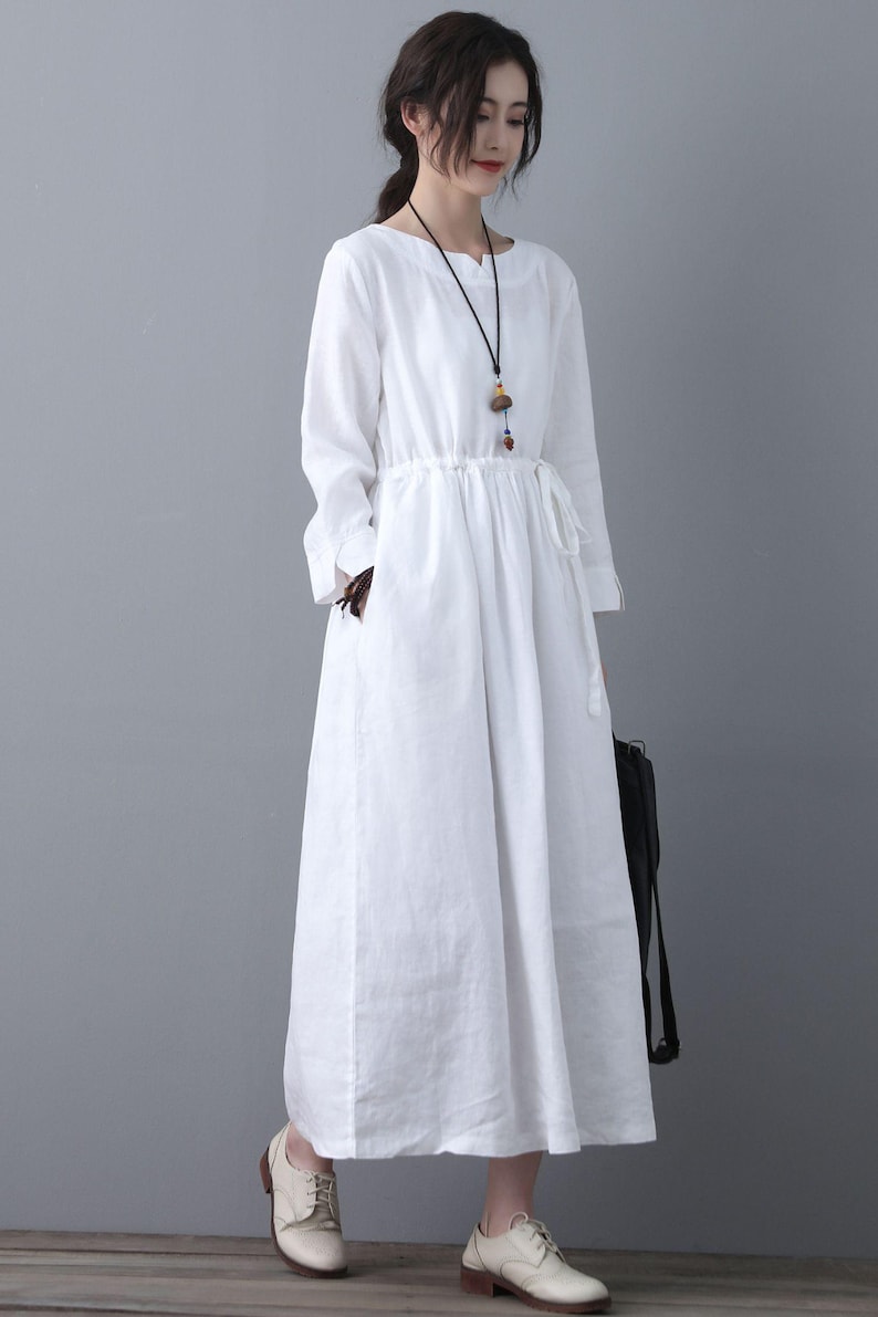 May include: A white linen dress with long sleeves and a drawstring waist. The dress has a relaxed fit and a slightly flared skirt. The model is wearing the dress with a simple necklace and white shoes.