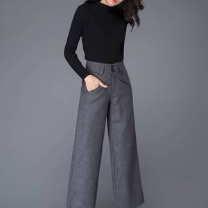Puede incluir: Una mujer con un su&eacute;ter negro de cuello alto y pantalones grises de pierna ancha. Est&aacute; de pie sobre un fondo gris.
