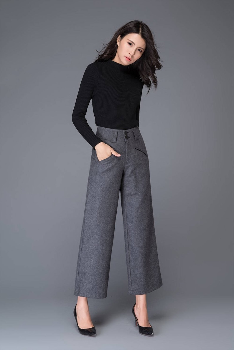 Puede incluir: Una mujer con un su&eacute;ter negro de cuello alto y pantalones grises de pierna ancha. Est&aacute; de pie sobre un fondo gris.