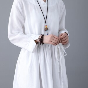May include: White linen dress with long sleeves and a tie at the waist. The dress has a relaxed fit and a slightly gathered skirt. The model is wearing a necklace with a wooden pendant.