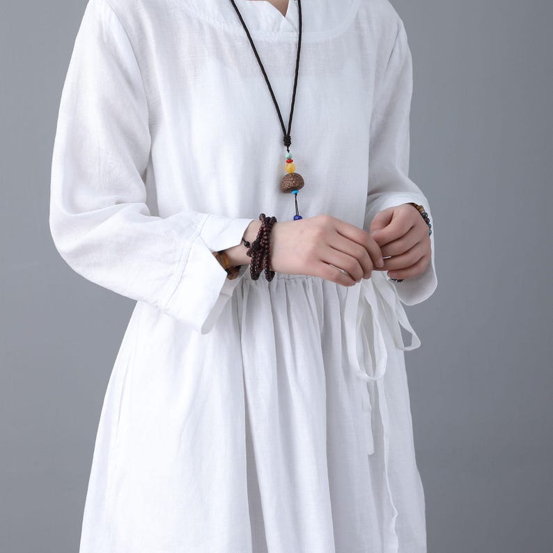 May include: White linen dress with long sleeves and a tie at the waist. The dress has a relaxed fit and a slightly gathered skirt. The model is wearing a necklace with a wooden pendant.