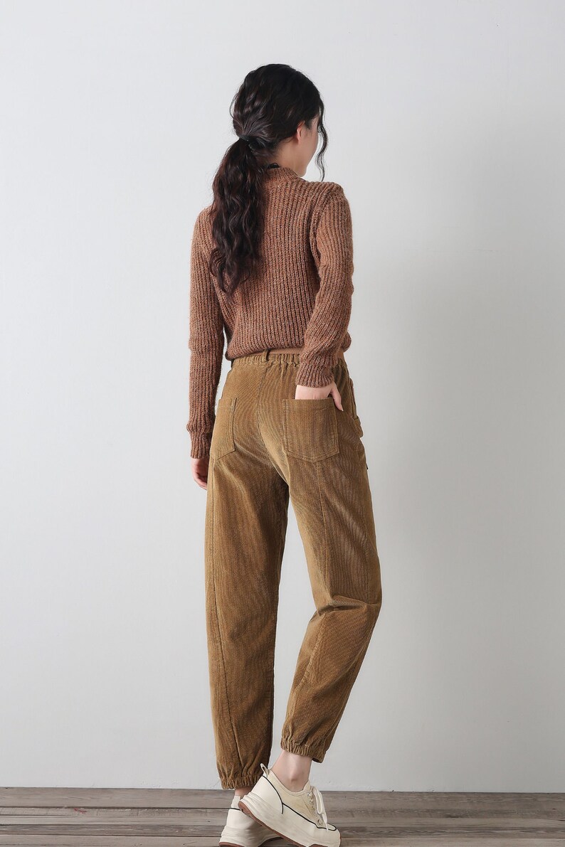 K&ouml;nnte beinhalten: Eine braune Cordhose mit lockerer Passform und elastischem Bund. Die Hose hat eine leicht verk&uuml;rzte L&auml;nge und wird an einem Model gezeigt, das einen braunen Pullover tr&auml;gt.