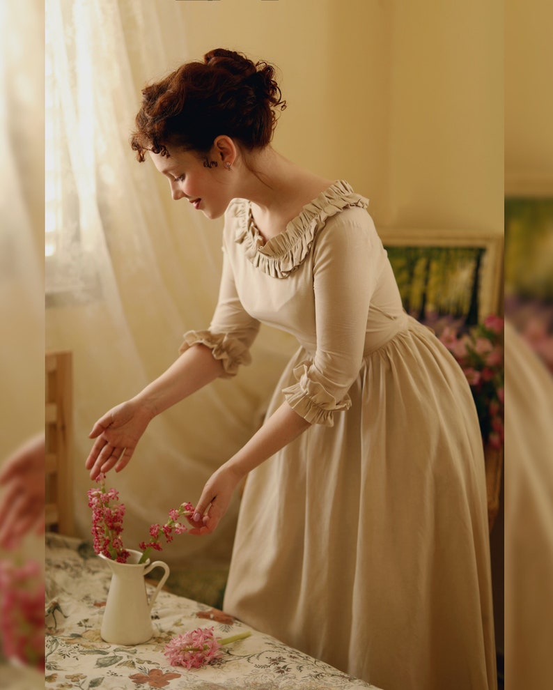 Puede incluir: Una mujer con un vestido beige con cuello y mangas con volantes est&aacute; arreglando flores rosas en una jarra blanca sobre un mantel floral.