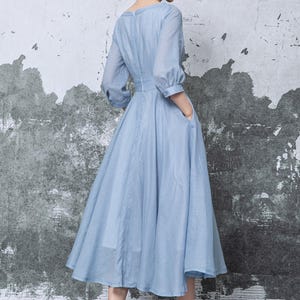 May include: A light blue dress with a V-neckline and elbow-length sleeves. The dress has a fitted waist and a flowing skirt that falls to mid-calf. The dress features pockets and is paired with white heels.