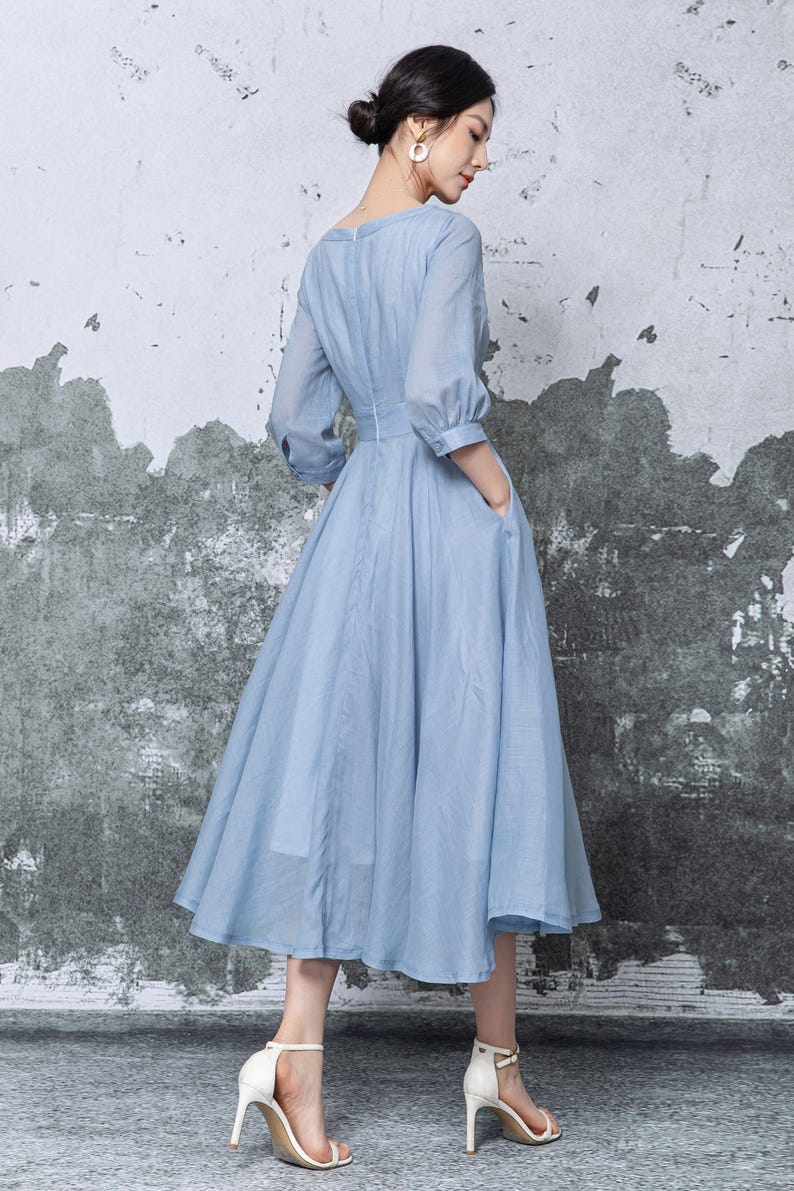 May include: A light blue dress with a V-neckline and elbow-length sleeves. The dress has a fitted waist and a flowing skirt that falls to mid-calf. The dress features pockets and is paired with white heels.