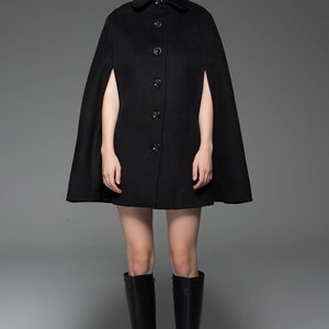May include: A black wool cape with a button-up front and long sleeves. The cape has a wide, flowing design and is worn over a black dress. The model is wearing black knee-high boots.
