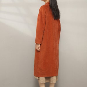 May include: A long, rust-colored corduroy coat with a collar and long sleeves. The coat is worn over a beige skirt and brown ankle boots.