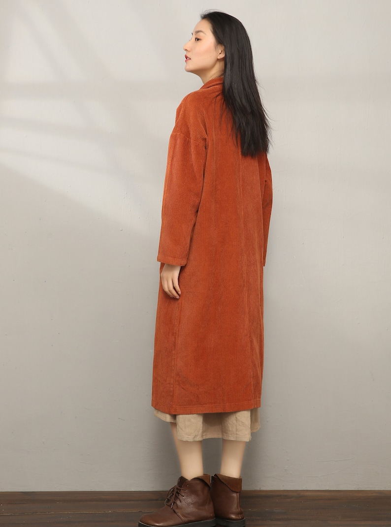 May include: A long, rust-colored corduroy coat with a collar and long sleeves. The coat is worn over a beige skirt and brown ankle boots.
