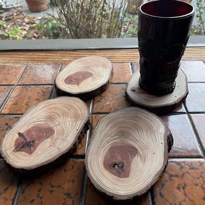 May include: Four natural wood coasters with a reddish-brown bark edge and a light wood center with brown rings. The coasters are arranged in a semi-circle with a dark red glass on top of one of the coasters.