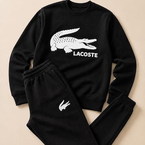 Op de afbeelding: Een zwart sweatshirt met lange mouwen en ronde hals en een bijpassende zwarte joggingbroek. Het sweatshirt heeft een grote witte krokodilafbeelding en de tekst LACOSTE. De broek heeft een kleiner wit krokodillogo op de linkerdij.