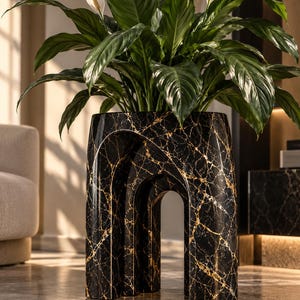 Pode incluir: Um vaso de mármore preto com veios dourados apresentando um design estrutural com duas aberturas em arco. O vaso contém uma grande planta de lírio-da-paz verde com duas flores brancas que se estendem da folhagem escura e brilhante.