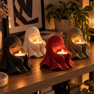 Peut inclure: Quatre bougeoirs en forme de fantôme de couleur noire, blanche, rouge et vert sauge. Chaque figurine a les yeux fermés et tient une petite bougie chauffe-plat allumée. Les sculptures mates et lisses sont alignées sur une surface en bois.