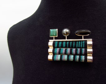 May include: A gold-toned brooch with a black, green, and gray striped fabric design. The brooch features a green square stone, a black oval stone, and a gray round stone.