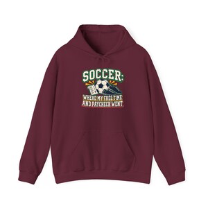 May include: A maroon long-sleeve hooded sweatshirt featuring a graphic print of a soccer ball, a receipt, and cleats. The text reads: SOCCER: WHERE MY FREE TIME AND PAYCHECK WENT. The design uses green, white, yellow, and black colors on the front.