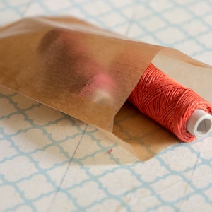 May include: A spool of bright orange thread is wrapped in a brown paper bag. The bag is lying on a white surface with a blue and white patterned design.
