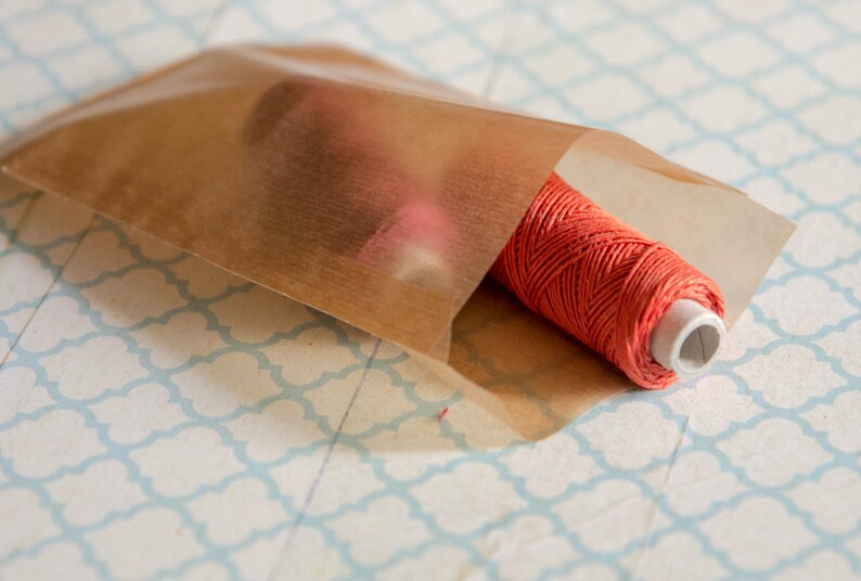 May include: A spool of bright orange thread is wrapped in a brown paper bag. The bag is lying on a white surface with a blue and white patterned design.
