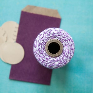 May include: A roll of white and purple twine with a brown cardboard spool. The twine is on a purple gift bag with brown paper backing. There are four round brown paper gift tags in the background.