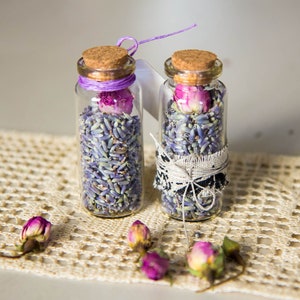 Puede incluir: Dos pequeñas botellas de vidrio llenas de lavanda seca y un solo capullo de rosa seco, atadas con una cinta y encaje. Las botellas están sobre un encaje de ganchillo.