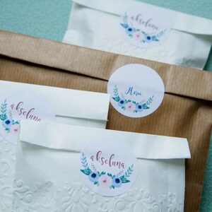 May include: Three white paper bags with floral stickers. The stickers have the names "akseluna" and "Mezu" written on them.