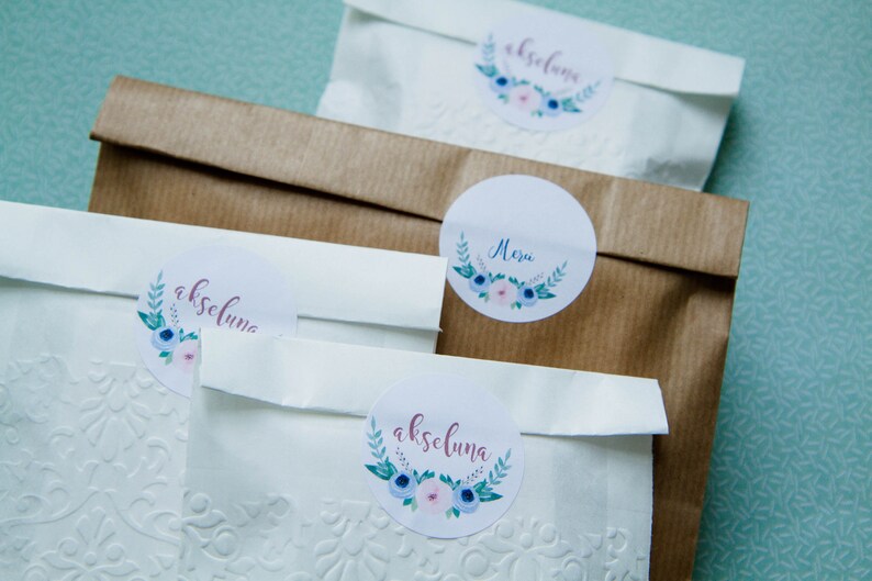 May include: Three white paper bags with floral stickers. The stickers have the names "akseluna" and "Mezu" written on them.