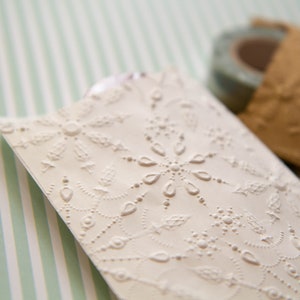 May include: A white paper gift bag with a delicate embossed pattern of a snowflake and leaves. The bag is on a striped green and white background.