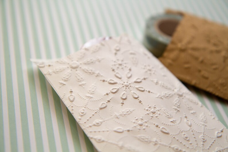 May include: A white paper gift bag with a delicate embossed pattern of a snowflake and leaves. The bag is on a striped green and white background.