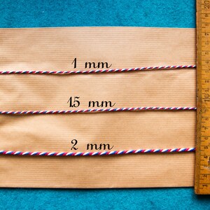 Pu&ograve; includere: Tre lunghezze di spago rosso, bianco e blu sono disposte su una superficie di carta marrone accanto a un righello di legno. Lo spago &egrave; etichettato 1 mm, 15 mm e 2 mm.