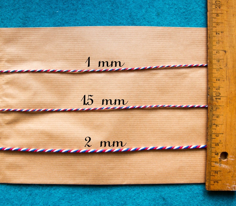Pu&ograve; includere: Tre lunghezze di spago rosso, bianco e blu sono disposte su una superficie di carta marrone accanto a un righello di legno. Lo spago &egrave; etichettato 1 mm, 15 mm e 2 mm.