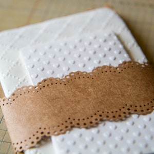 May include: Two white cardstock envelopes with a brown paper band and a white lace trim. The envelopes have a raised pattern of small hearts.