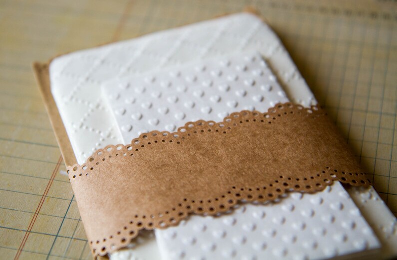 May include: Two white cardstock envelopes with a brown paper band and a white lace trim. The envelopes have a raised pattern of small hearts.