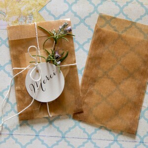 May include: Two brown paper bags, one tied with twine and a white tag that says "Merci", and the other is empty. Both bags are on a blue and white patterned surface.