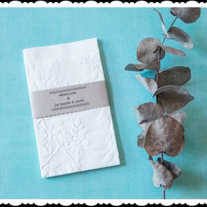May include: White envelope with a floral embossed design and a brown label that reads "ackeluna & la bande a part". The envelope is on a light blue background with a dried eucalyptus branch.