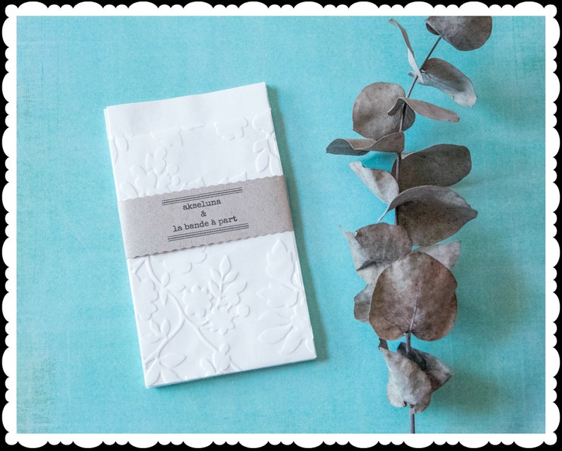 May include: White envelope with a floral embossed design and a brown label that reads "ackeluna & la bande a part". The envelope is on a light blue background with a dried eucalyptus branch.