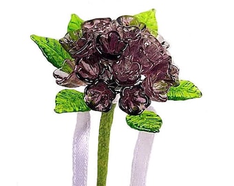 May include: A glass bouquet of purple flowers with green leaves and white ribbons.