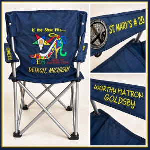 May include: A navy blue folding chair with a silver metal frame. The back features an embroidered Order of the Eastern Star logo, a high-heeled shoe, and the text If the Shoe Fits, OES, Order of Eastern Star, and Detroit, Michigan. Armrests display St. Mary's #20 and Worthy Matron Goldsby.