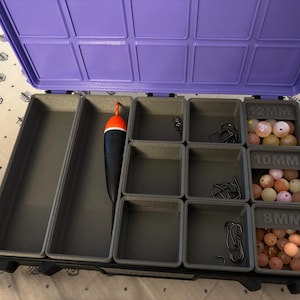May include: A purple plastic tackle box with a hinged lid containing gray modular compartments. The interior holds a black and orange fishing float, metal hooks, and three labeled bins filled with round beads marked 12mm, 10mm, and 8mm.