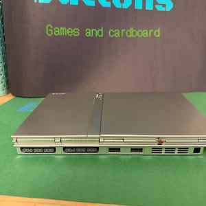 May include: A silver-colored game console featuring a rectangular design with two memory card slots, two controller ports, and two USB ports on the front panel. The console is positioned in front of a dark sign with the text Buttons Games and cardboard.
