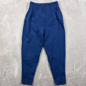 May include: High-waisted blue trousers featuring a structured waistband with two dark buttons, front pleats, and reinforced stitching panels on the inner legs. These pants have a tapered leg silhouette and side pockets, providing a clean, functional design.
