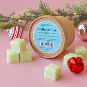 May include: A close-up of a brown container with a label that reads "Mistletoe Kiss sugar scrub cubes." Several light green cubes are scattered around the container, along with red and silver Christmas ornaments. The product is a weekly, moisturizing body exfoliant.