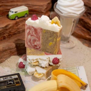 May include: Stacked handmade soaps resembling a dessert. The soaps have layers of pink, brown, and white, decorated with raspberries and banana slices. A green and white toy van and a sundae glass are in the background.