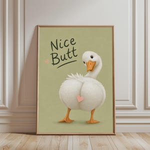 Op de afbeelding: Een ingelijste kunstdruk met een witte eend met een oranje snavel die over de schouder terugkijkt. De achtergrond is gedempt olijfgroen. De tekst Nice Butt is in het zwart gedrukt, vergezeld van twee kleine roze hartjesillustraties.