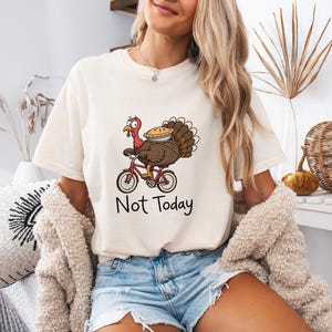 May include: A cream-colored short-sleeve t-shirt featuring a graphic of a cartoon turkey riding a red bicycle and carrying a pie on its back. Below the graphic, the text "Not Today" is printed in a simple black sans-serif font.