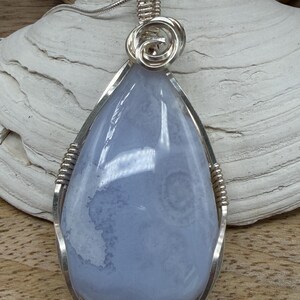 May include: A teardrop-shaped blue chalcedony pendant encased in a polished silver wire-wrapped setting. The stone features soft, cloudy patterns in shades of light blue and white. A silver chain attaches to a decorative coiled wire bail at the top.
