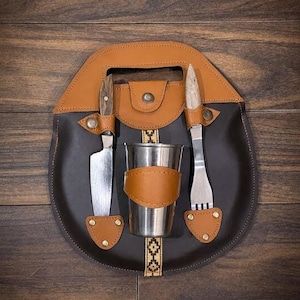 May include: A dark brown leather carrying case with a tan leather handle and trim, featuring a central stainless steel cup, a knife with a wooden handle, and a fork with a wooden handle, all secured by tan leather straps with brass-colored metal snaps.