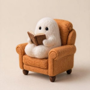 May include: A needle-felted white ghost figure with black bead eyes sits in a miniature orange armchair. The ghost holds a small, open brown book with visible pages, resting its arms on the book while seated in the textured fabric chair.